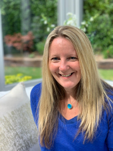 A smiling blonde woman with a blue top and a turquoise pendant necklace, photographed in a bright indoor setting with a blurred garden background.